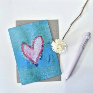 A greeting card, aqua coloured crayon background with a pink heart on the middle, the pink heart has a red crayon outline. It sits in front of an envelope, there is a pale pink pen beside and a white paper-daisy sitting partially on top of the card.